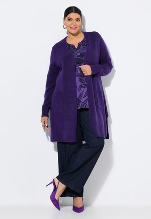 Violettes Strickcardigan mit zwei vorderen Taschen, getragen über einem gemusterten Oberteil und einer marineblauen Hose, kombiniert mit lila High Heels.