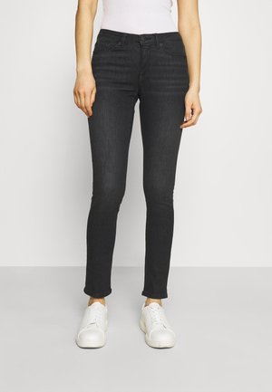 Femme portant un jean noir slim, des baskets blanches et un haut blanc, debout devant un fond clair uni.