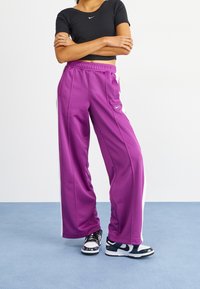 Fioletowe spodnie sportowe z białymi paskami bocznymi, elastycznym paskiem w talii i dwiema bocznymi kieszeniami, sparowane z czarnym dopasowanym topem i trampkami.