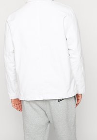 En person som bär en vit långärmad jacka och ljusgrå Nike sweatpants, visad bakifrån mot en enkel bakgrund.