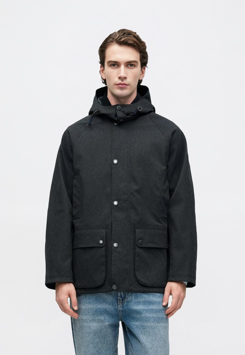 Young man wearing a dark gray hooded jacket with button front and large pockets, paired with light blue jeans, standing against a white background.