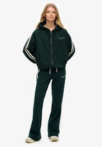 Dunkelgrüne Kapuzenjacke mit weißen Streifen und verstellbarer Kapuze, kombiniert mit passenden Jogginghosen. Beide Teile haben ein Logo.