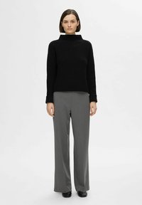 Pull à col roulé noir côtelé à manches longues, associé à un pantalon large gris taille haute, porté avec des chaussures noires. Fond uni.