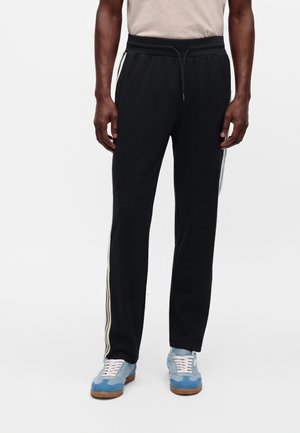 Uomo che indossa pantaloni da ginnastica neri con strisce laterali bianche e sneakers azzurro chiaro con lacci bianchi, in piedi davanti a uno sfondo semplice.