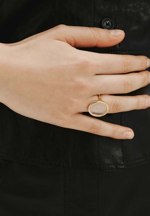 Mano descansando sobre tela negra que lleva un anillo de oro con una piedra oval translúcida grabada con un delicado diseño floral.