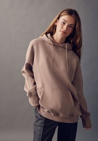 Bluza z kapturem w jasnym odcieniu taupe, z przednią kieszenią, luźnym krojem i ściągaczami na mankietach; zestawiona z ciemnymi spodniami skórzanymi.