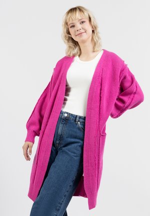 Lange roze cardigan met een ribbelstructuur, open voorkant en oversized mouwen. Draagt over een witte top en hooggetailleerde blauwe jeans.