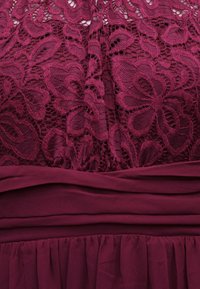 Σκούρο κόκκινο δαντελένιο ύφασμα με λουλουδένια σχέδια πάνω από στιβαρές στρώσεις υφάσματος με πτυχώσεις και σούρες.