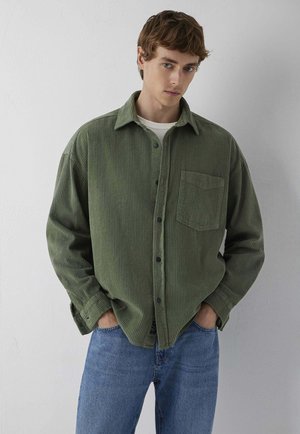Junger Mann, der ein olivgrünes Cordhemd über einem weißen Unterhemd und blauen Jeans trägt, mit den Händen in den Taschen vor einer schlichten Wand steht.