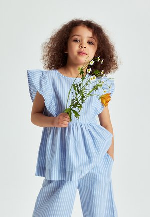 Nuori tyttö, jolla on kiharat hiukset, pukeutuneena siniraidalliseen mekkoon ja housuihin, pitää käsissään pientä luonnonkukkakimppua, johon kuuluu päivänkakkaroita ja oranssi kukka.