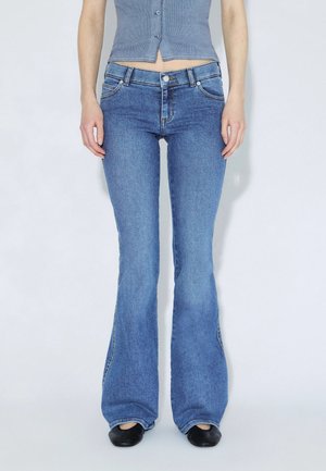 Personne portant un jean évasé bleu taille haute, un top gris court à boutons et des chaussures noires, debout devant un fond clair uni.