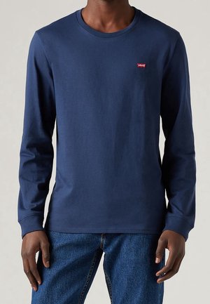 Personne portant un T-shirt manches longues bleu marine uni avec un petit logo rouge Levi's sur la poitrine gauche, associé à un jean bleu.