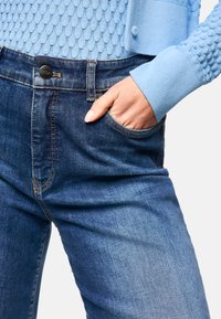 Dark blue denim jeans with a slim fit, featuring a front pocket, belt loops, and subtle stitching details. Textured blue knit top partially visible.
