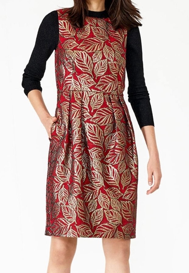 Robe rouge avec un motif de feuilles dorées et noires, corsage ajusté, jupe évasée et manches longues noires. Comprend des poches. Tissu texturé.