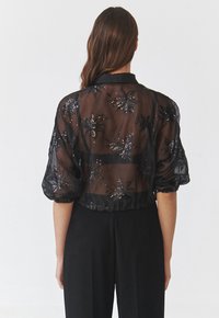 Bluză neagră din material transparent, cu mâneci bufante și aplicații din paiete florale. Design scurt cu tiv rotunjit și textură moale.