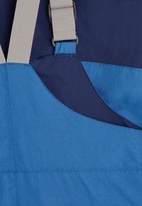 Blauw en marineblauw stoffen schort met beige banden, voorzien van overlappende delen en geaccentueerde stiksels, ontworpen voor functionaliteit en duurzaamheid.
