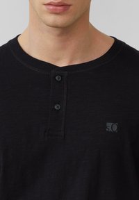 Crna kratka majica s kratkim rukavima tipa henley s tri gumba, okruglim izrezom i suptilnom teksturom tkanine. Ima mali logo na lijevoj strani.