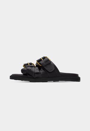 Μαύρα σανδάλια με σαγιονάρα, με δύο φαρδιές λουρίδες, διακοσμημένες με αγκράφες σε χρυσό τόνο και ρυθμιζόμενα κλεισίματα σε επίπεδη σόλα, επάνω σε απλό φόντο.