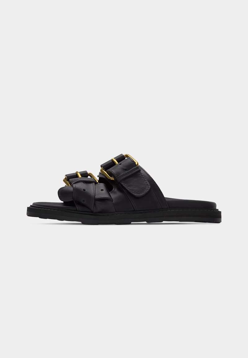 Μαύρα σανδάλια με σαγιονάρα, με δύο φαρδιές λουρίδες, διακοσμημένες με αγκράφες σε χρυσό τόνο και ρυθμιζόμενα κλεισίματα σε επίπεδη σόλα, επάνω σε απλό φόντο.