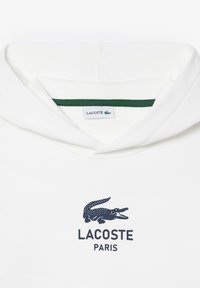 Felpa bianca con cappuccio, caratterizzata da un colletto interno verde, con un logo di coccodrillo navy e la scritta "LACOSTE PARIS" sottostante. Tessuto morbido, vestibilità standard.