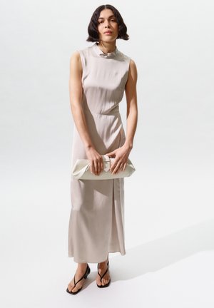 Femme aux cheveux courts et foncés portant une robe midi beige clair sans manches, tenant une pochette blanche et des sandales noires à brides, debout sur un sol blanc.