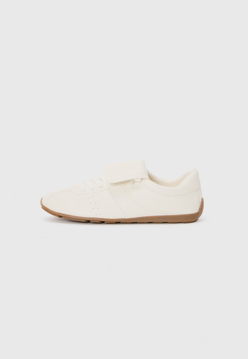 Biała sneakers z gładką cholewką, z sznurówkami i zapięciem na rzep, oraz brązową gumową podeszwą. Posiada perforowane akcenty na bokach.
