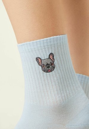 Calzino azzurro chiaro a coste su una gamba con una faccia di bulldog francese grigia ricamata vicino alla caviglia.