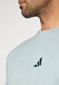 Κοντινό πλάνο ενός άνδρα που φοράει ανοιχτό μπλε πουκάμισο με ένα μικρό σκούρο πράσινο στιλιζαρισμένο λογότυπο στο στήθος σε απλή φόντο.