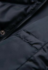 Marinblå vadderad jacka med slätt, glänsande tyg, dragkedjestängning och en fickdetalj på vänster ärm.