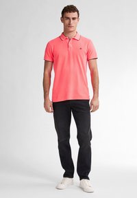 Polo shirt corail avec un col, des manches courtes et des bords noirs contrastants, associé à un jean foncé et des baskets blanches.