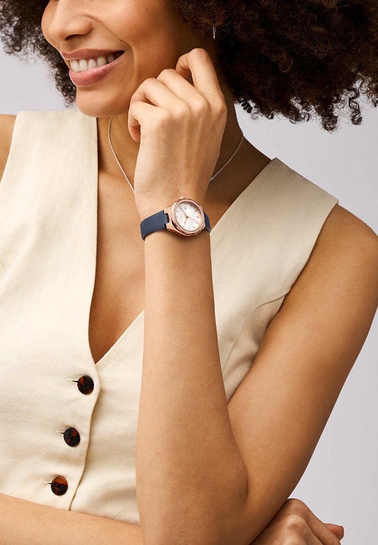 Reloj de oro rosa con esfera blanca y correa de silicona azul marino, con una caja redonda y marcadores de hora sutiles. Botones destacados en el lateral.