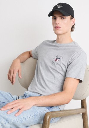Camiseta de algodón gris con cuello redondo y logo de GANT, combinada con jeans de mezclilla azul y una gorra negra con texto blanco, sentado en una silla clara.