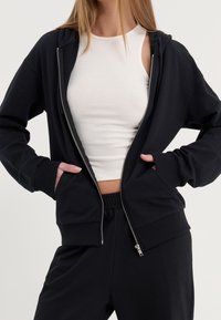 Schwarzer Kapuzenpullover mit Reißverschluss, glatter Textur, gerippten Bündchen und Kängurutasche. Trägt ein weißes geripptes Tanktop darunter.