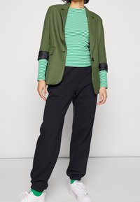 Personne portant un blazer à carreaux verts et noirs sur une chemise à manches longues à rayures vertes et blanches, un pantalon noir, des chaussettes vertes et des baskets blanches.