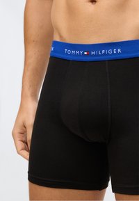 Torso masculino vestindo boxers pretos com uma faixa azul rotulada "Tommy Hilfiger" e um pequeno logotipo da marca.