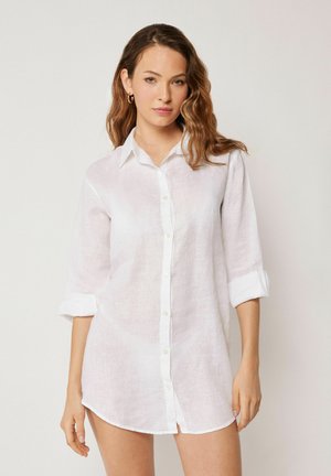 Kvinde med bølget hår iført en lang, hvid, knap-op-skjorte med op-rullede ærmer, stående mod en ensartet lys baggrund.