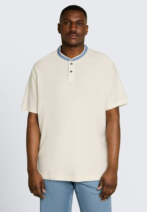 Man staand, vooruitkijkend, draagt een crèmekleurig kort mouwen overhemd met een blauwe knoopsluiting aan de kraag en lichtblauwe broek.