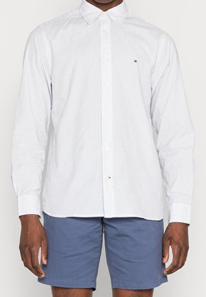 Mann trägt weißes, langärmliges Hemd mit dünnen vertikalen Streifen und blaue Shorts, steht mit entspannten Armen seitlich am Körper.