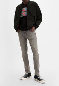 Black bomber jacket over a dark graphic t-shirt, paired with light grey jeans and black high-top sneakers with white soles.