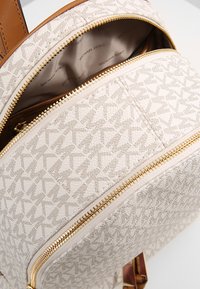 Beige textuur rugzak met een geometrisch patroon, gouden ritsen en een bruine leren schouderband. Het interieur is voorzien van een logoprint.