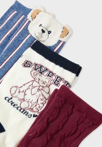 Tre paia di calzini colorati per bambini, con un motivo a righe blu e la faccia di un orsetto, un paio bianco con un orsetto e la scritta "sweet dreams", e un paio bordeaux con dettagli in maglia.