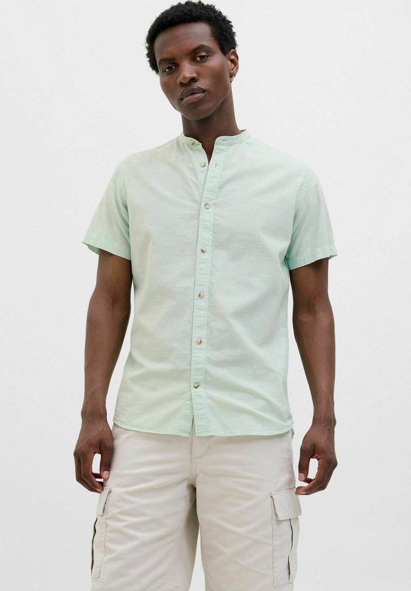 Camicetta verde chiaro a maniche corte con colletto alla francese, chiusura a bottoni sul davanti e tessuto testurizzato. Il modello indossa pantaloni cargo beige.