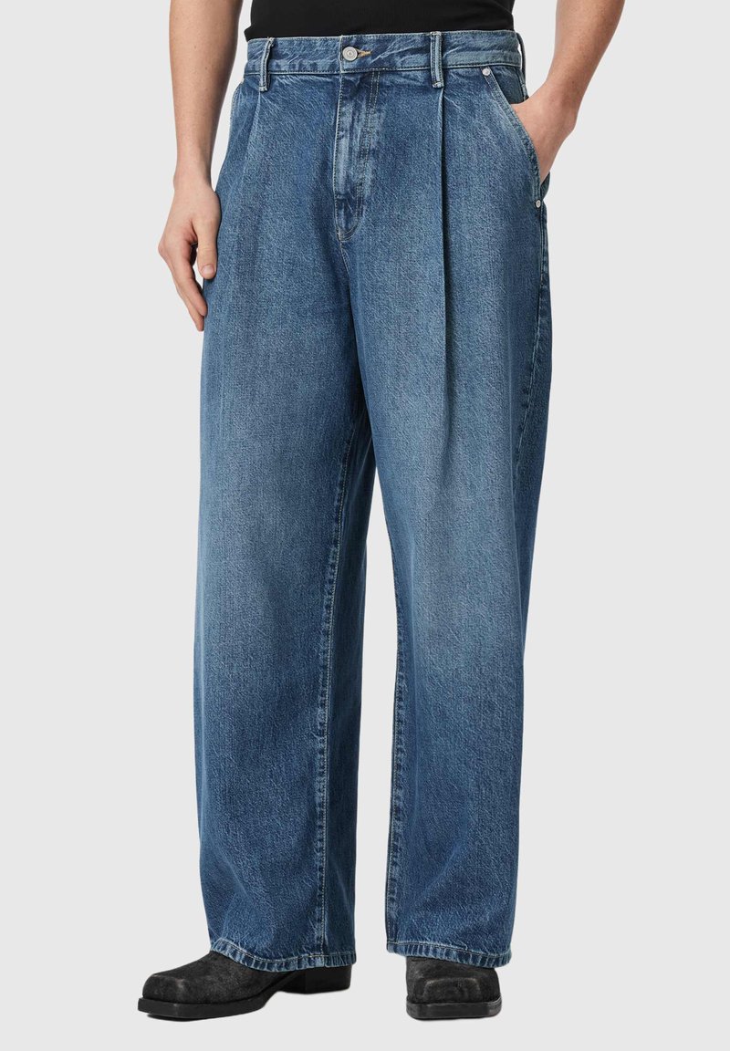 Personne portant un jean denim bleu ample avec des plis, des poches avant et des chaussures noires, debout devant un fond clair uni.