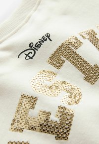 Fehér pulóver arany fényes felirattal és Disney logóval. Az anyaga puha, bordázott nyakkal és sima textúrával rendelkezik.