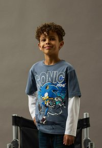 Leende pojke med lockigt hår som bär en blå långärmad Sonic Prime-tröja, stående framför en neutral grå bakgrund och en svart stol.