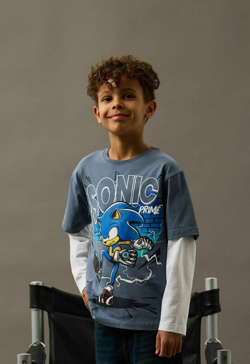 Leende pojke med lockigt hår som bär en blå långärmad Sonic Prime-tröja, stående framför en neutral grå bakgrund och en svart stol.