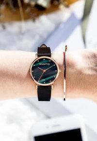 Czarny skórzany zegarek na pasku z okrągłą różowozłotą kopertą, zieloną tarczą w paski i kryształowymi znacznikami godzin. Obok znajduje się cienka różowozłota bransoleta.