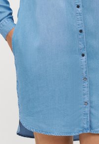 Jasnoniebieska sukienka koszulowa z długimi rękawami, z przodem na guziki, bocznymi kieszeniami i zaokrąglonym wykończeniem. Gładka tekstura i klasyczny design.