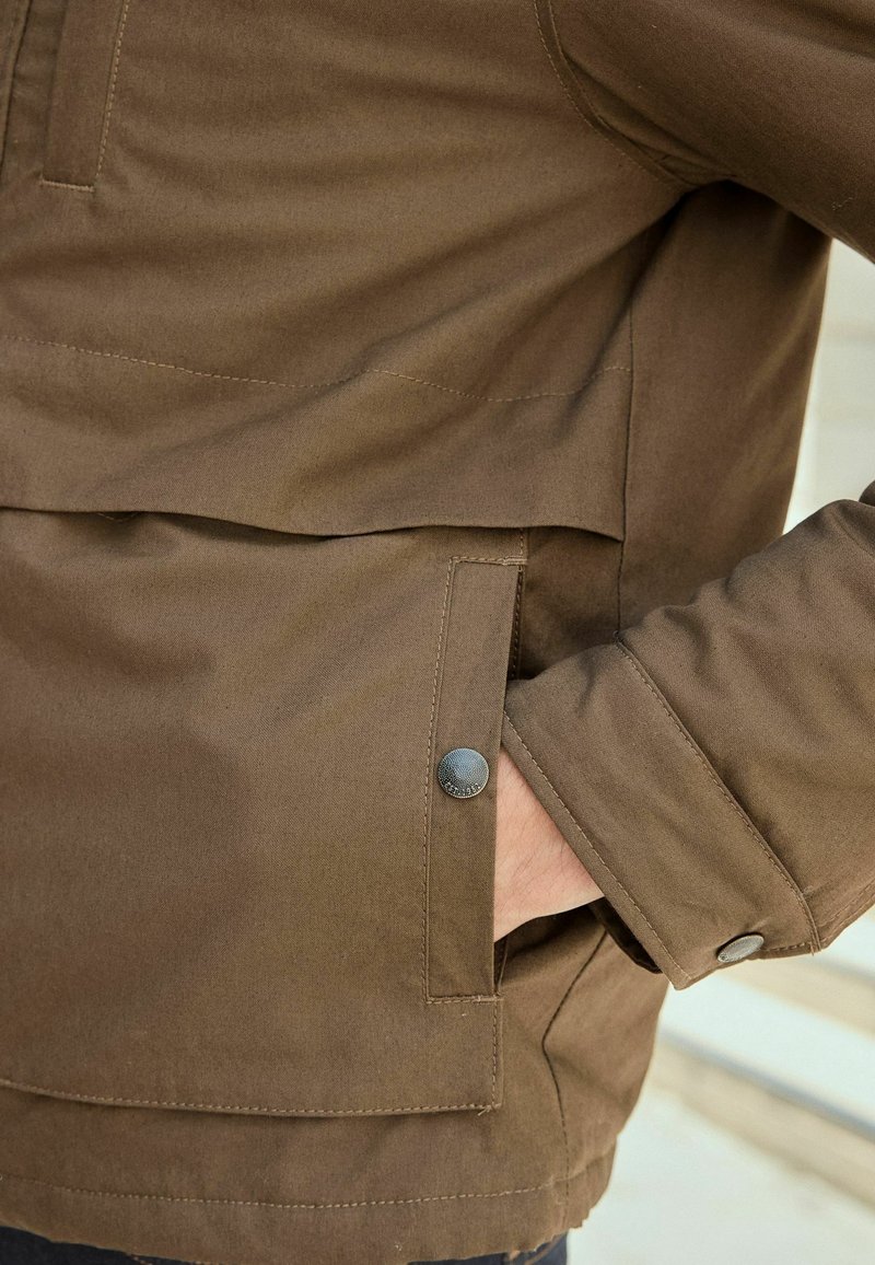 Gros plan sur la manche d'une veste marron et une poche avant avec un bouton-pression, une main partiellement à l'intérieur de la poche.