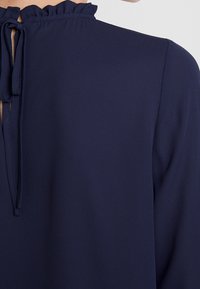 Blouse bleu marine avec un col à volants et un dos à trou doublé de liens, fabriquée dans un tissu lisse à légère brillance.